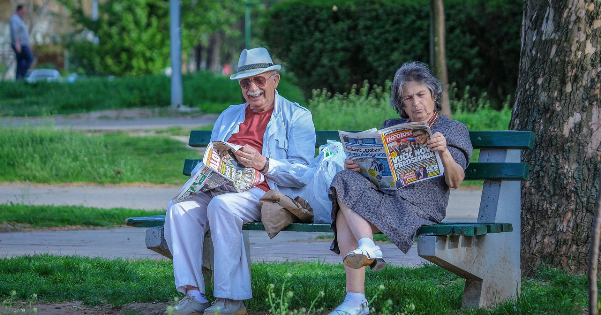 Two elderly couples sitting on a bench, reading newspapers, reflecting the importance of wellness and mental well-being in later years