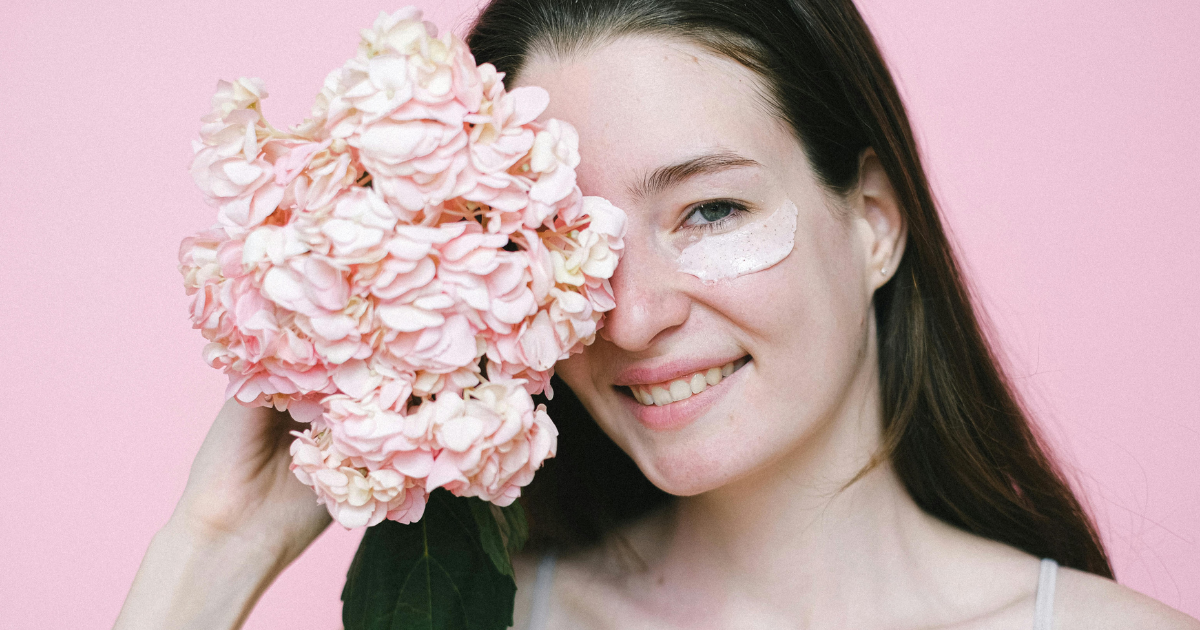 A woman pampering herself with an under-eye mask and holding a bunch of flowers, illustrating the role of self-care.