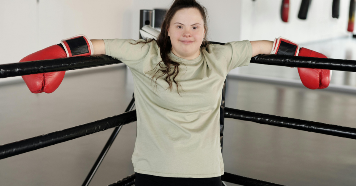 A teenage girl with Down syndrome leans against a boxing ring with gloves, challenging societal norms and proving that self-transformation is possible despite unique challenges.