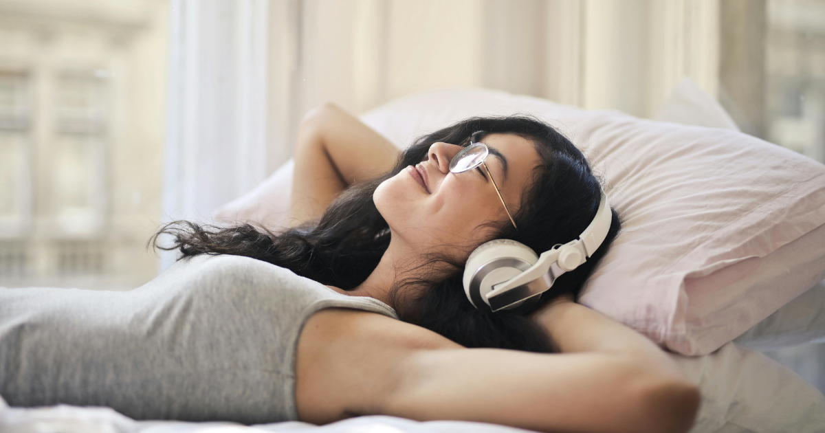 Image: A woman lying down with closed eyes, listening to music through headphones, portraying the joy of letting go of the past and embracing the present moment."