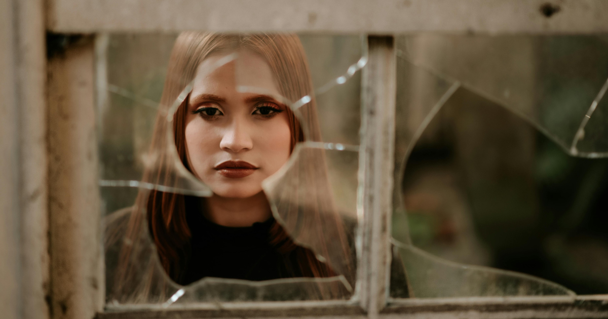 Image: An intense woman gazes at shattered window glass, symbolizing the significance of breaking rules metaphorically.