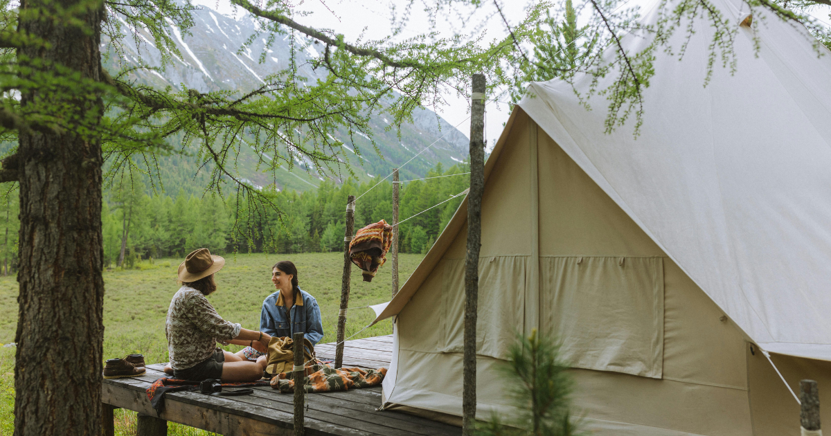 Image: Two individuals sit outside a tent house in a secluded forest area, defying the norms of materialistic society.