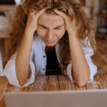 Image description: A woman sits in front of her laptop, holding her head tightly with both hands, signifying stress induced by online trolling.