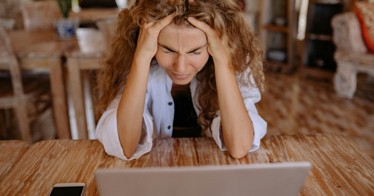 Image description: A woman sits in front of her laptop, holding her head tightly with both hands, signifying stress induced by online trolling.