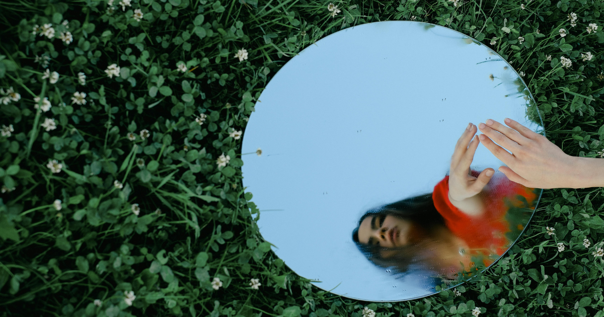 A woman touches her reflection in a mirror on the ground, symbolizing introspective self-talk.