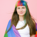 A young woman wearing a rainbow flag, standing and smiling, shows the boldness of being from the LGBTQ community.