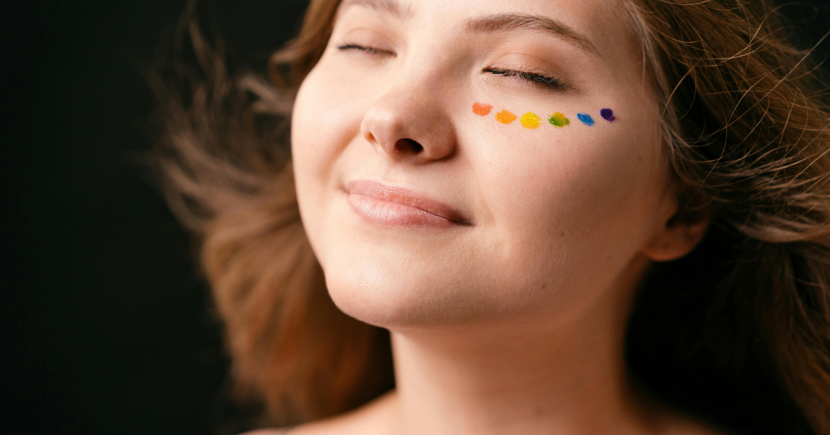 A young woman with closed eyes and a smile, with rainbow paint below her eyes, shows self-acceptance and love as an LGBTQ individual.
